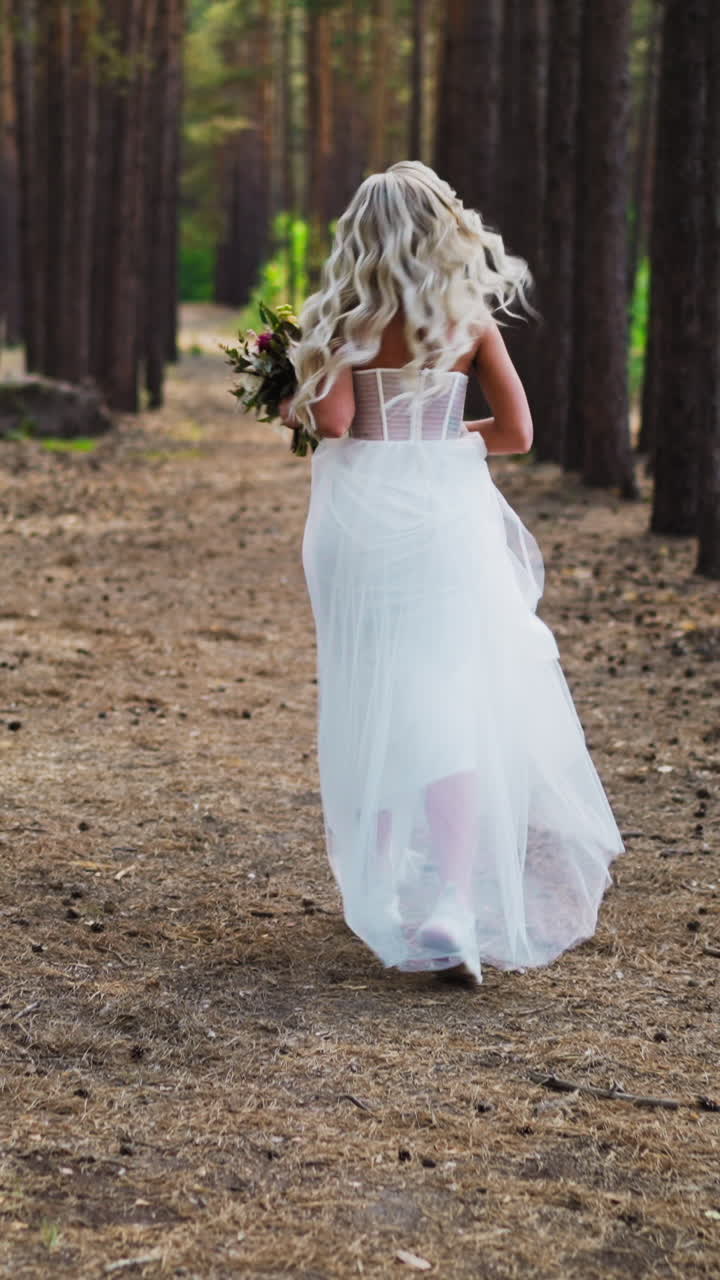 Pretty young woman with curly locks in stylish white wedding dress and sneakers runs and turns head back along pine forest backside view slow motion