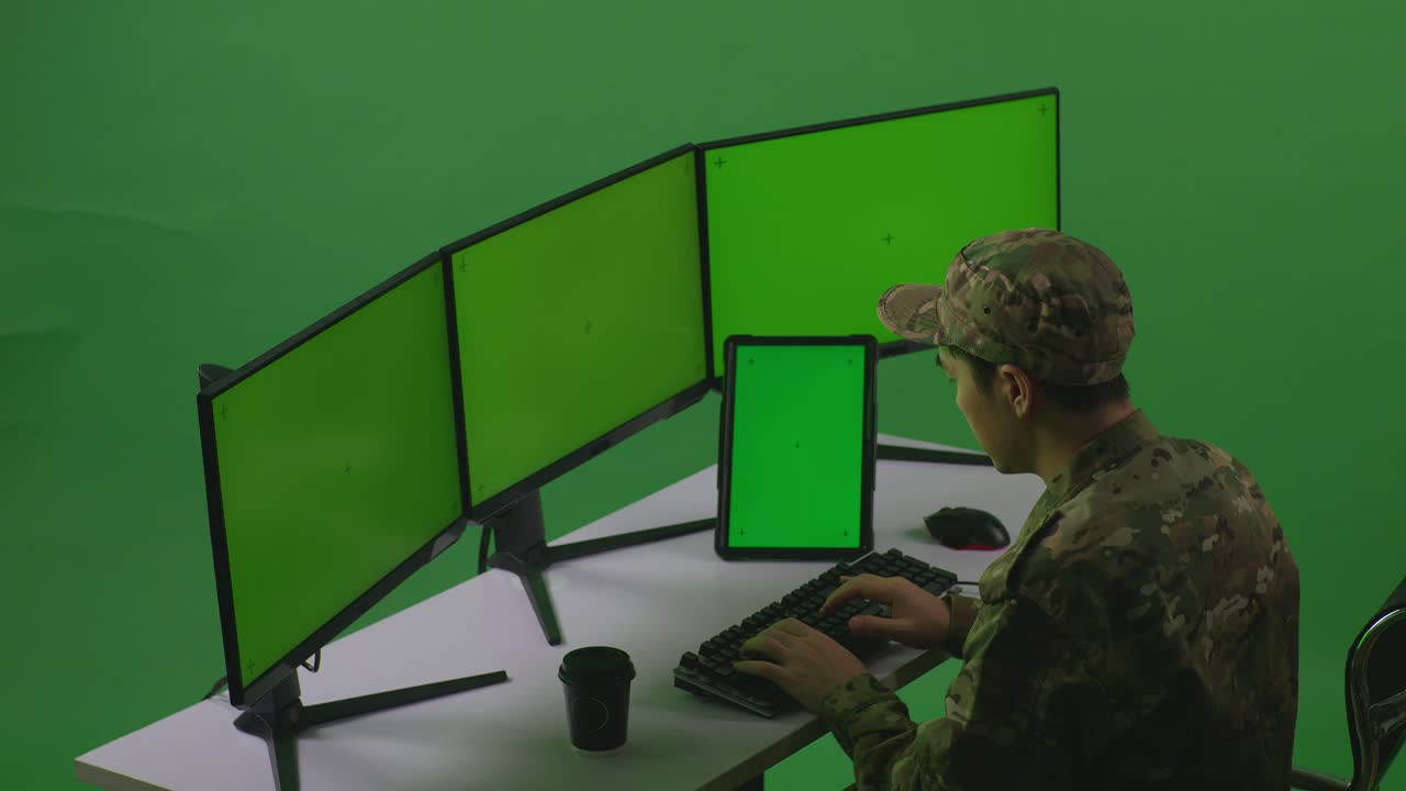 боковой вид азиатского мужчины-солдата, работающего с планшетом и несколькими компьютерными мониторами с макетным зеленым экраном в студии зелёного экрана