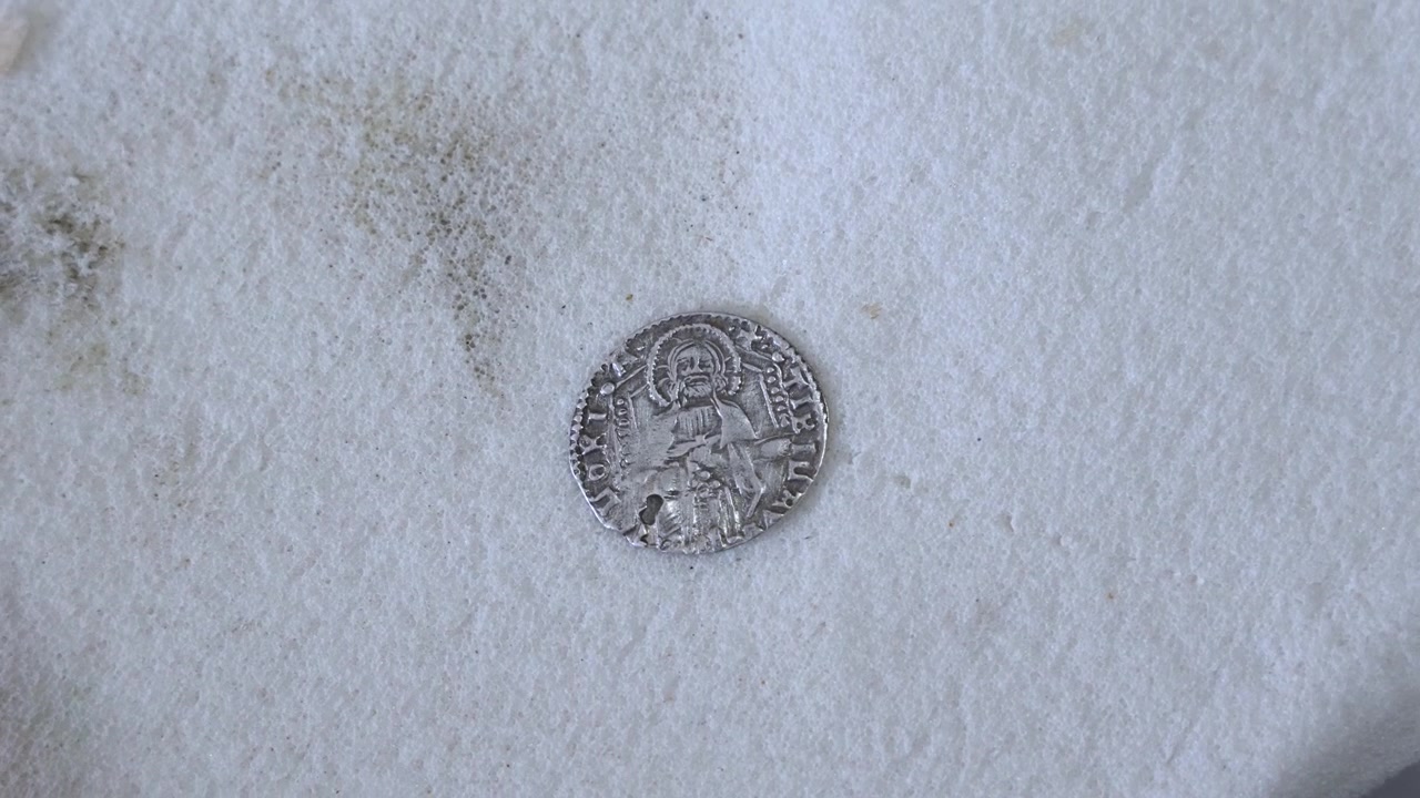 Primo piano di un'antica moneta d'argento ritrovata sotto il Castello di Shobak, Giordania, che mostra una figura con aureola, lettere consumate e una coniatura in stile medievale invecchiato su una superficie bianca