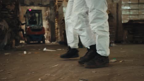 Close-up-shot-of-two-engineer-employees-in-white-protective-uniforms-and-black-shoes-walking-along-a-plant-for-recycling-waste-paper-and-plastic