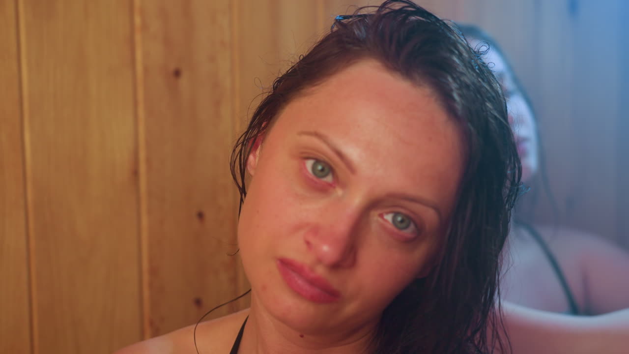 two women wrapped in towels relax in smoky sauna, one gazing upward with gentle smile under warm amber steam glow, damp hair framing face against wooden wall