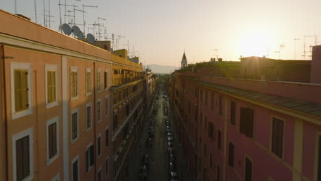 Forwards-fly-along-top-of-apartment-houses-in-street-of-urban-borough-at-sunrise.-Revealing-silhouette-of-tower-against-bright-sky.-Rome,-Italy