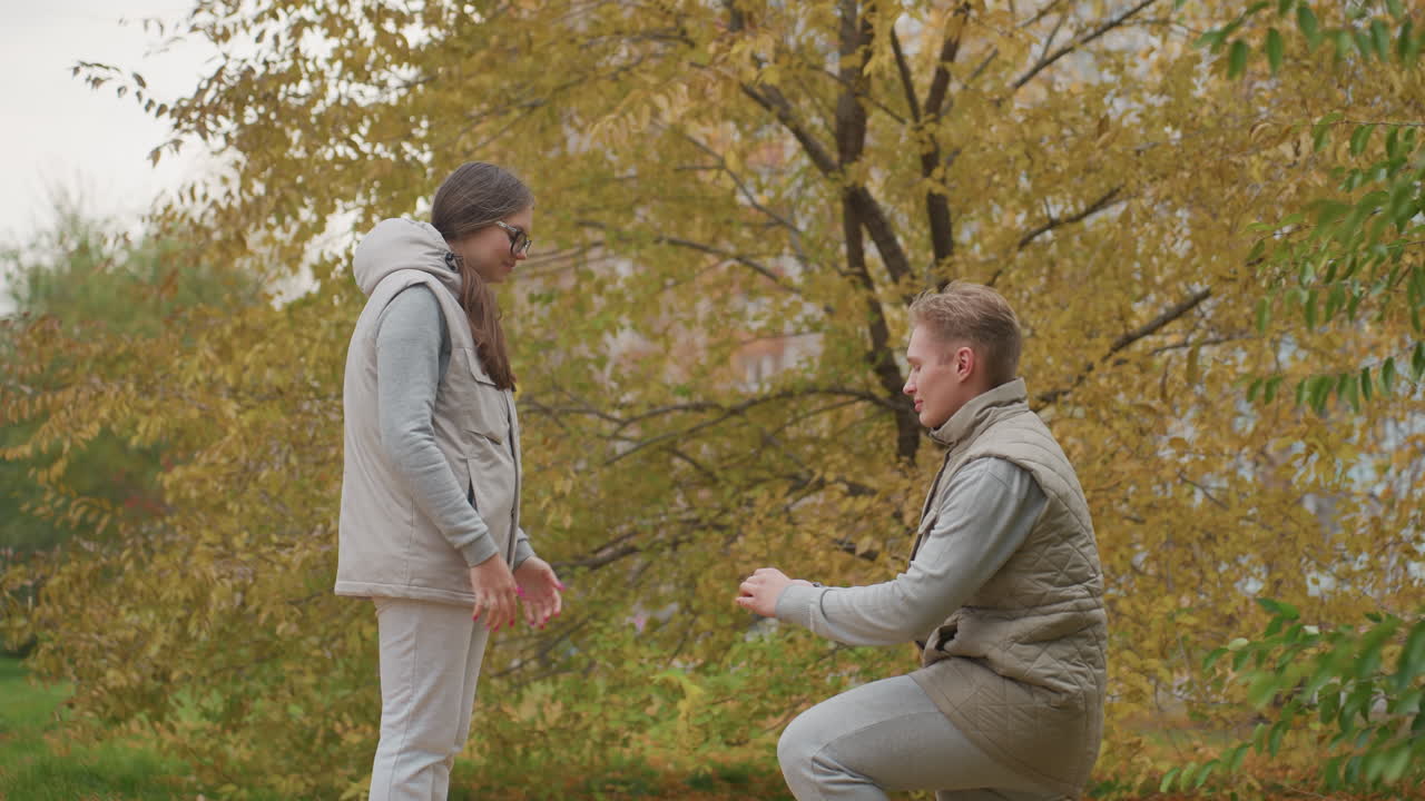 Young adult kneels down to present engagement ring to his love in romantic outdoor park setting filled with autumn leaves, while she reacts with joy and surprise, background