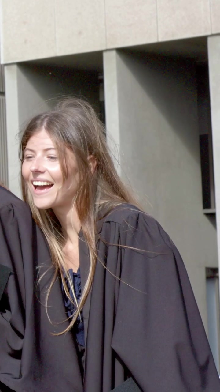 Video verticale: Laureato sorridente in toga si gira verso i colleghi che arrivano nel campus, guardando il telefono e il diploma.