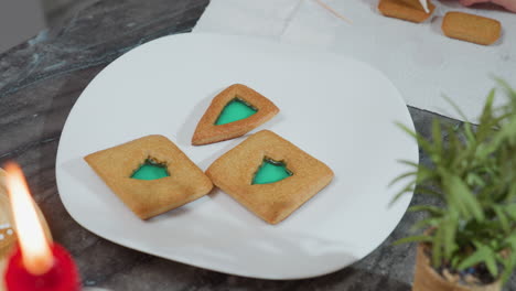 tres galletas decoradas en una placa blanca con relleno verde, vela roja ardiente y flor decorativa en la mesa, otras dos galletas colocadas en papel de textura blanca