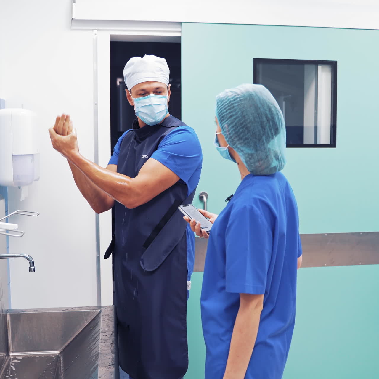 Surgeons in medical uniform and masks on their faces washing hands at the hospital. Doctors man and a woman talking while the man is washing his hands before the operation in clinic.