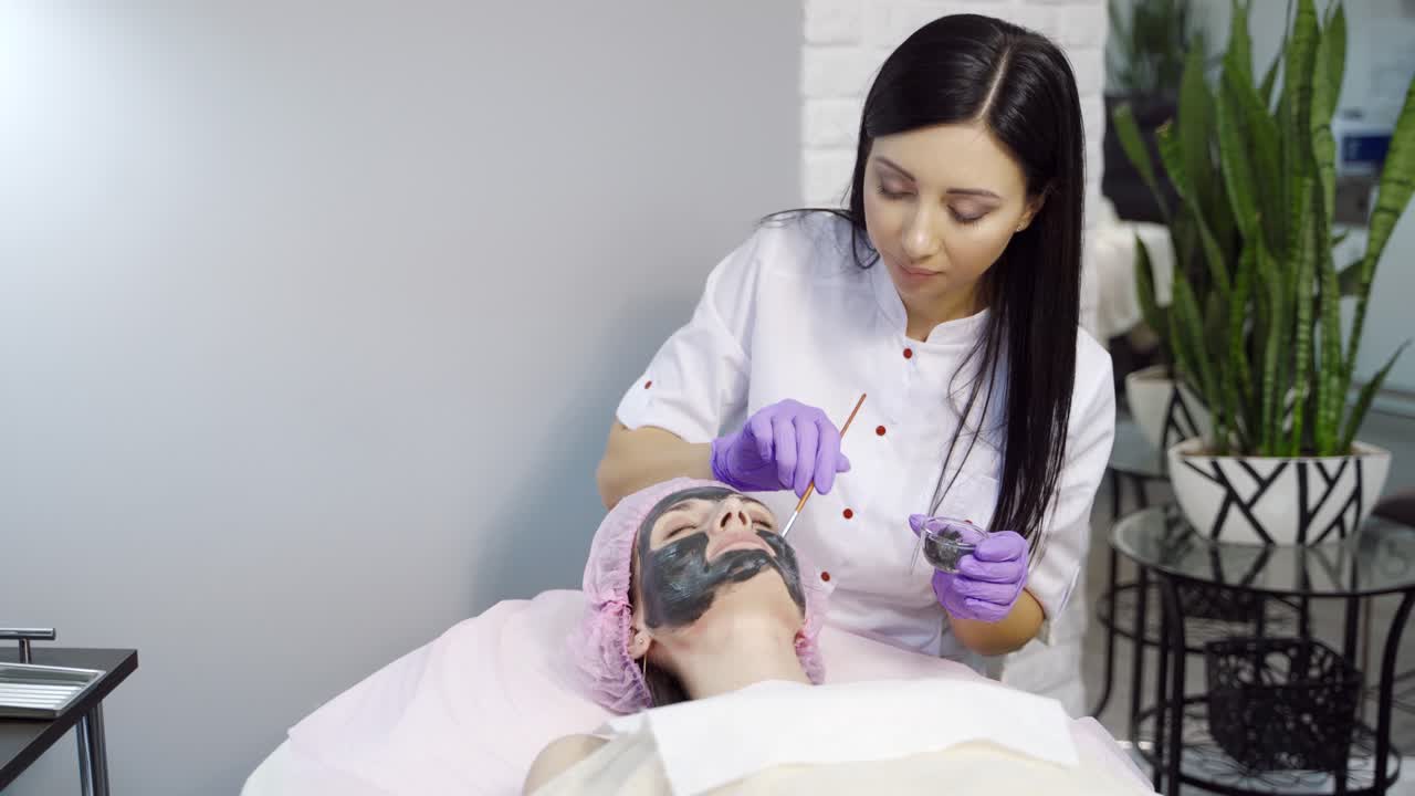 Pretty cosmetologist with long hair makes a mask to a woman for cleansing the pores on the face in cosmetology. Cosmetic procedures. Spa