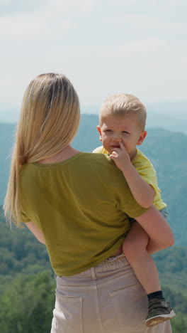 loving mother kisses cute toddler boy in arms standing against large mountains silhouettes at eco resort in summer backside view slow motion