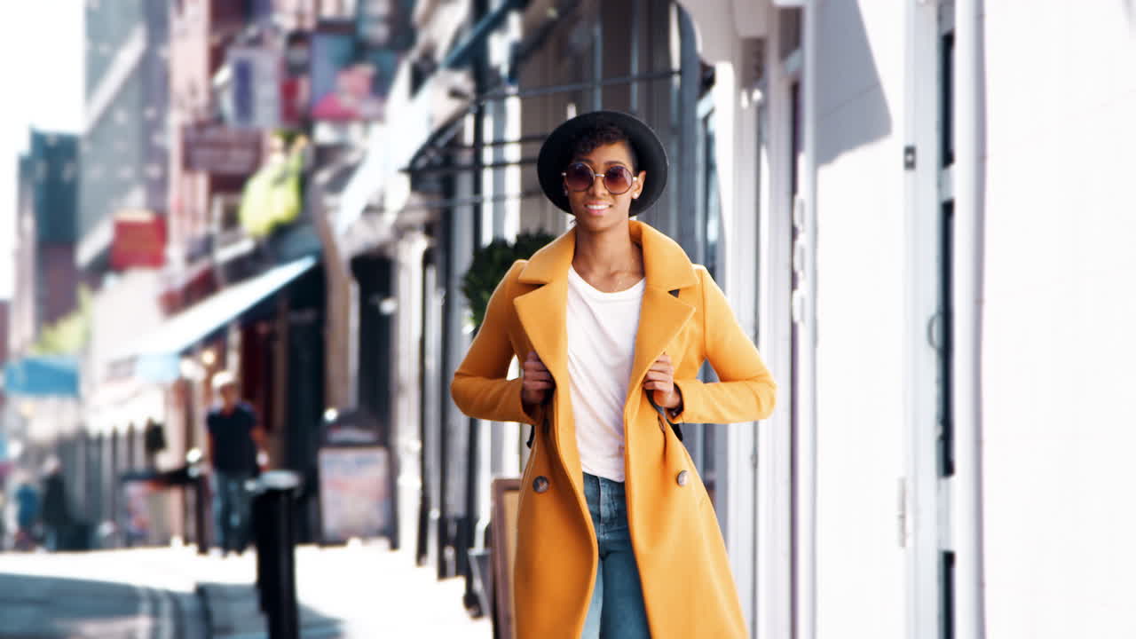 Fashionable young black woman wearing blue jeans and an unbuttoned yellow pea coat walking on pavement near shops on a sunny day smiling, close up