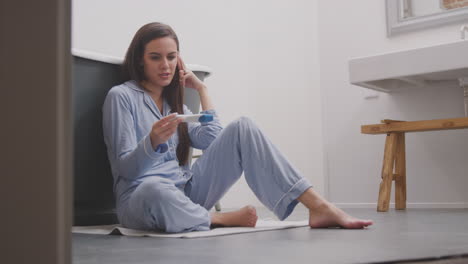 excited woman sitting on bathroom floor with positive home pregnancy test