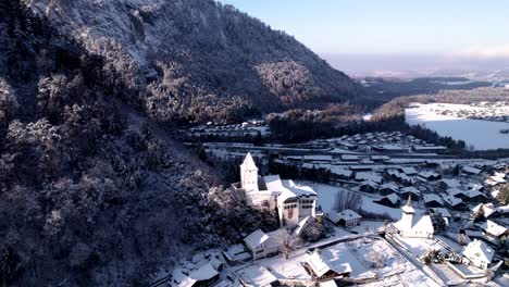 Luftaufnahme-Eines-Bergdorfes-Mit-Einer-Kirche-Und-Schneebedeckten-Felsigen-Bergen-Im-Hintergrund-In-Der-Schweiz-An-Einem-Sonnigen-Wintertag