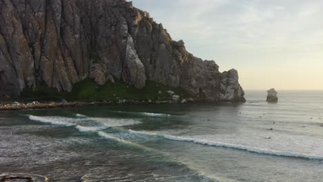 wide-aerial-of-surfers-in-the-ocean-trying-to-catch-waves-in-the-Pacific-Ocean-at-Morro-Bay-Rock-Beach-in-California-USA-during-sunset