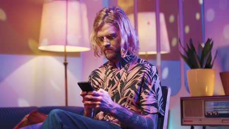 young stylish caucasian man with long hair in glasses sitting on a chair and using smartphone in a retro party at home