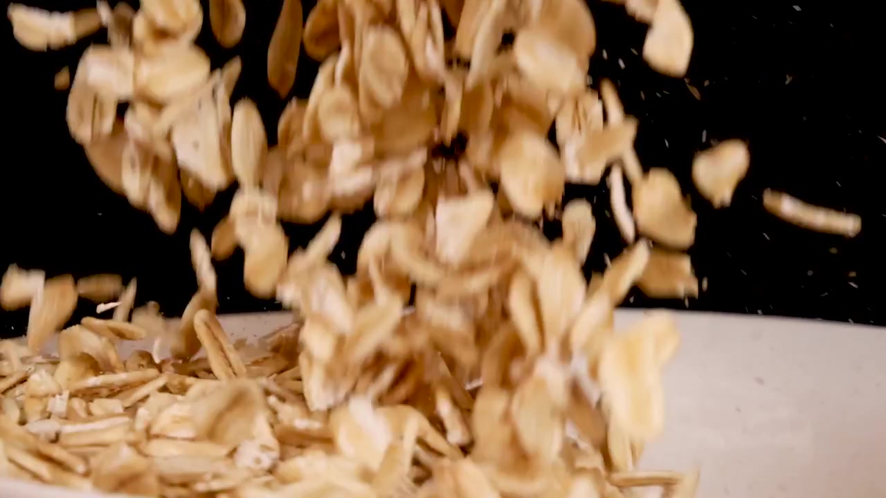 Primo piano di fiocchi d'avena biologici che cadono a cascata in una ciotola su sfondo nero.