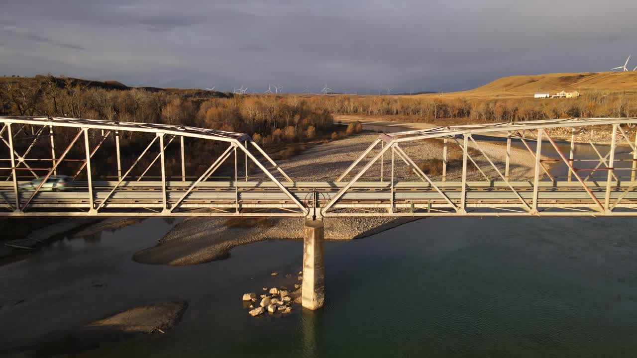 Slowly backwards flying drone revealing a beautiful old steel bridge in remote Alberta, Canada during sunrise. Dark cloudy sky over wind turbines in background. Car crossing the old man river in 4k.