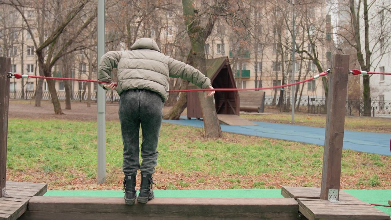 Person balancing on wooden beam in park during cold autumn day, hooded puffer jacket, slow cautious steps over worn platform, bare trees and fallen leaves, red barrier tape and distant playground