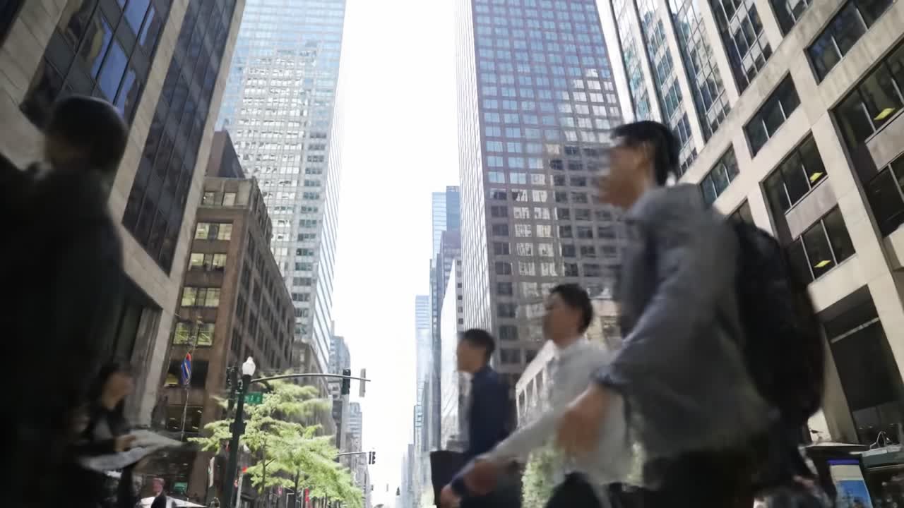 Pedestrians navigate a bustling city street lined with towering skyscrapers under a bright sky, showcasing the lively urban atmosphere at midday.