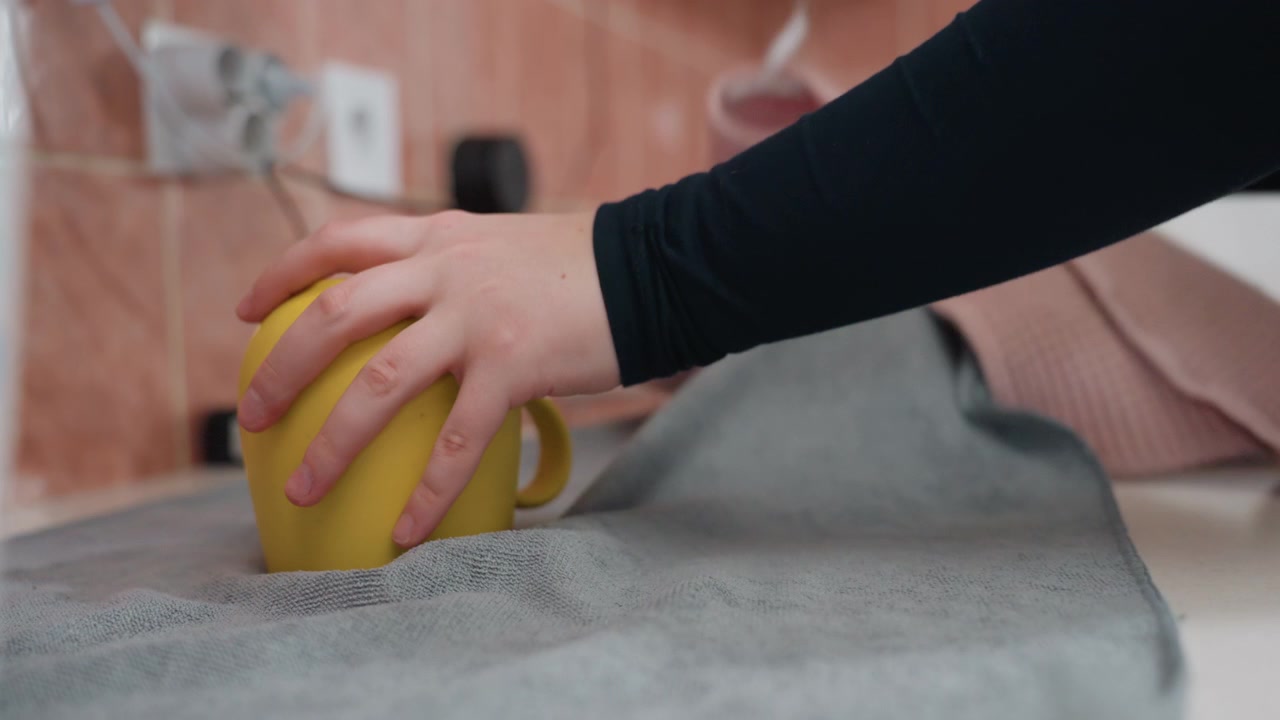 Taza amarilla sobre una toalla, una mano levanta la taza en la cocina de un pequeño apartamento, gestos finales de limpieza y empaque antes de la mudanza, persona destapa y recoge la taza dejando una marca húmeda en la toalla, primer plano
