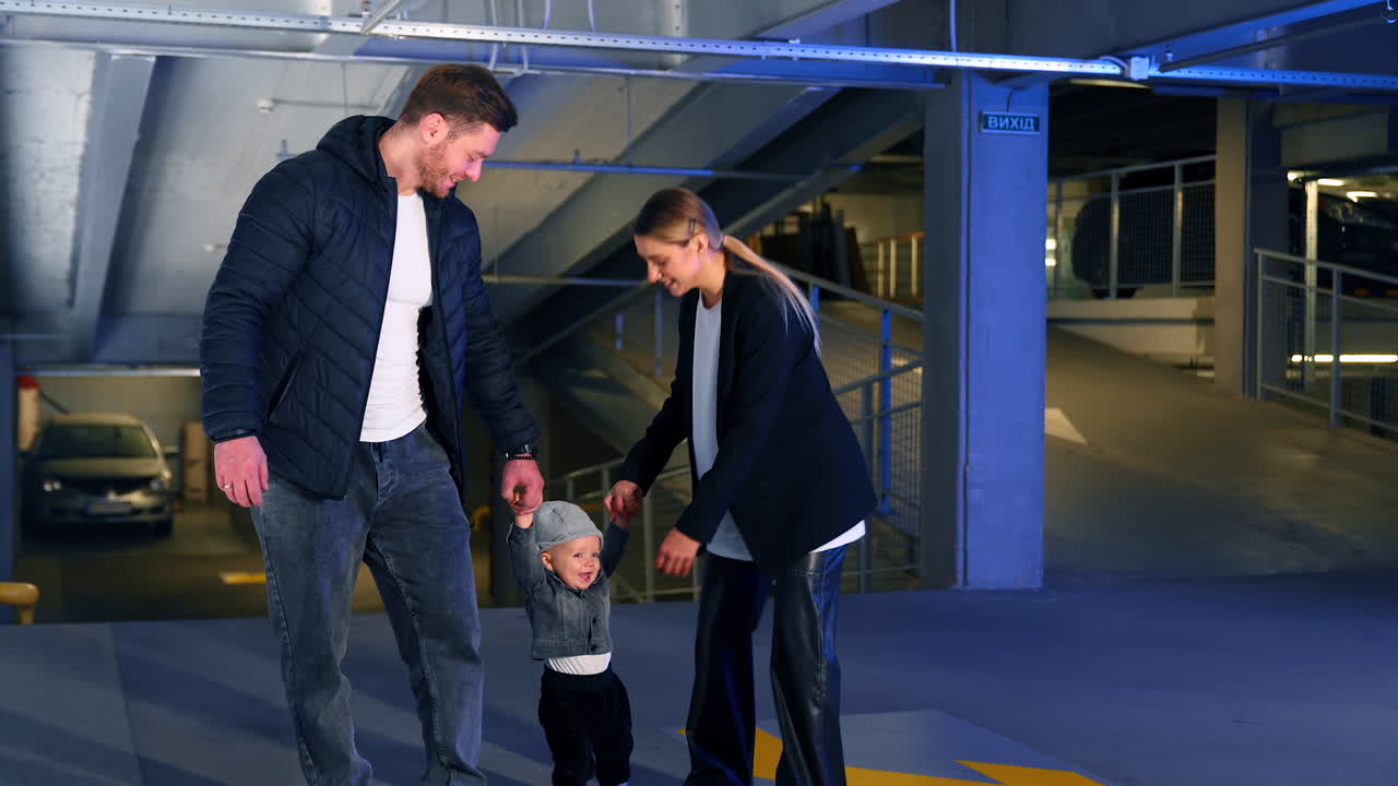 Parents walk holding their little baby by hands. Mother takes her child and turns around with him cheerfully.