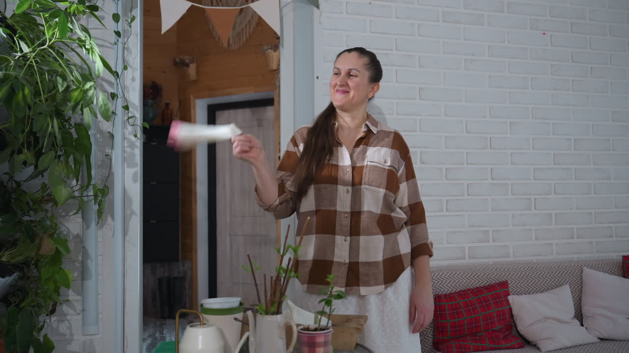 Plant lover smiles warmly while rolling napkin in hands next to glass table filled with potted greenery eyes focused on vibrant leaves home setting with brick wall natural light pouring from window