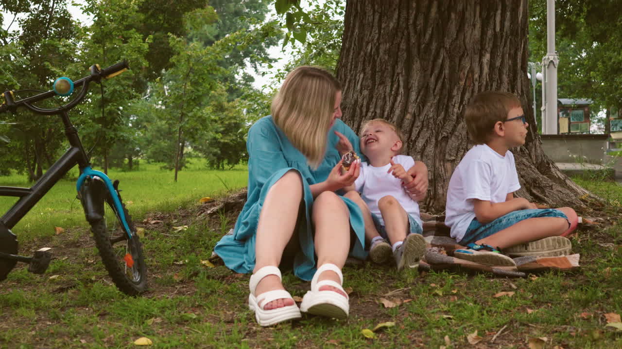 una madre sostiene a su hijo menor felizmente mientras le ofrece un pastel, ambos sonriendo de alegría, el niño mayor mira hacia otro lado, aparentemente molesto