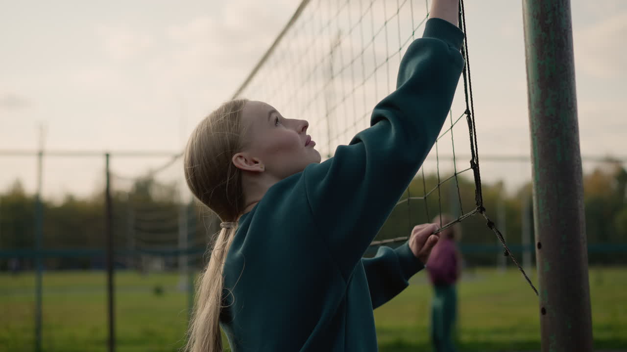 Close-up of young lady in green sweater checking firmness of volleyball net with blurred view of someone playing volleyball in the background during an outdoor sports training session