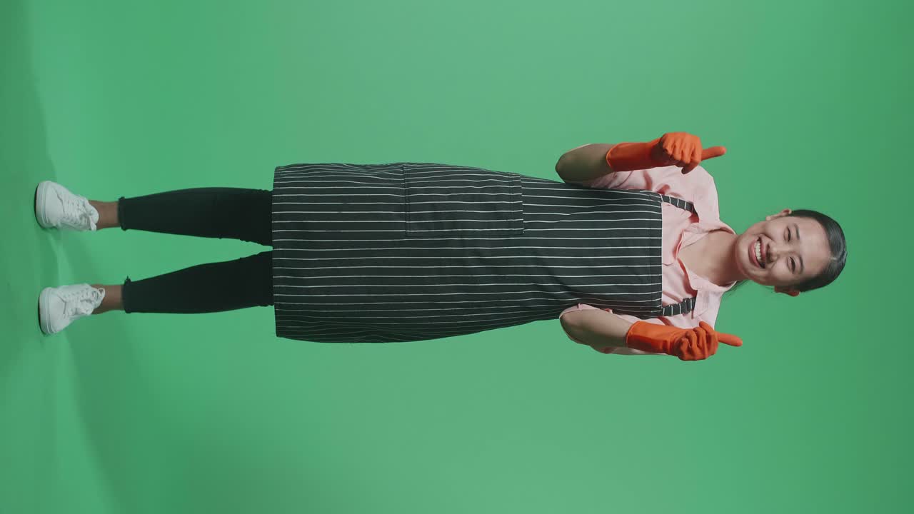 초록색 스크린 스튜디오에 서 있는 동안 미소 짓고 엄지손가락을 올리는 몸짓을 하는 아시아 여성 집사.