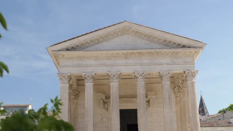 behind branches and green leaves, an imposing roman building facing forward with light-coloured stone columns, blue sky