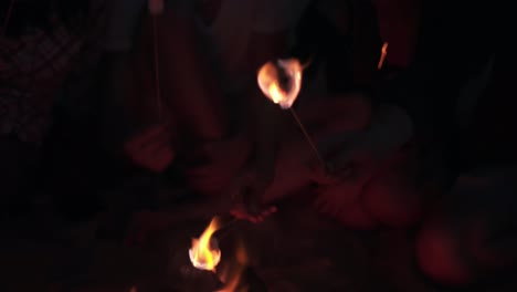 young cheerful friends sitting by the fire on the beach in the evening, cooking marshmallow on sticks together. shot in 4k