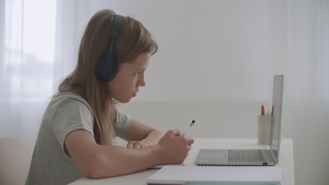 una estudiante está haciendo la tarea sentada en la mesa en su habitación usando una computadora portátil y auriculares escribiendo en un libro de ejercicios