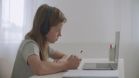 schoolgirl is doing homework sitting at table in her room using laptop and headphones writing in exercise book