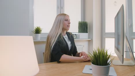 tired woman waking up at workplace. tired blond female office worker in elegant suit relaxing lying on arms on desk then getting up yawning and starting working on computer in light room with plants.