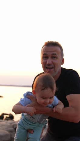 father playing with little son at the shore. cute baby boy is waved in dad's hands closer to camera. clear sky at backdrop. vertical video
