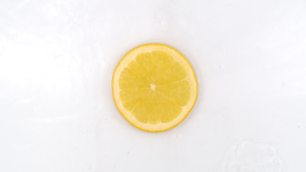 Top view: on a white background a fresh orange is lying in the water water drops are falling from above and splashes are falling in slow motion in all directions.