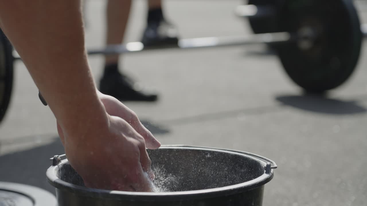 Close-up of young man puts chalk on his hands from bucket and claps so the air fills with dust before lifting wait at cross fit outside in 4k slow motion