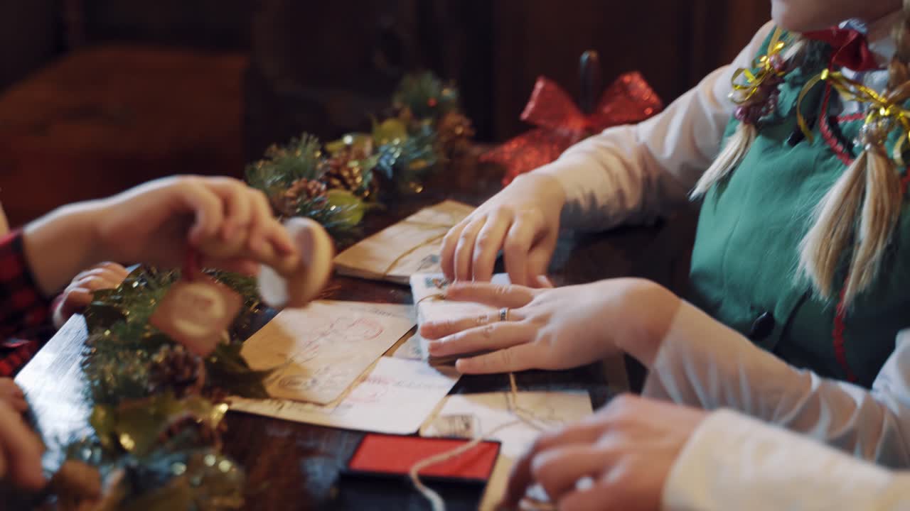 Elves and children making envelopes at Christmas. Many Christmas letters on the table and cute kid puts seal on the envelope.Female elf ties stack of envelopes.