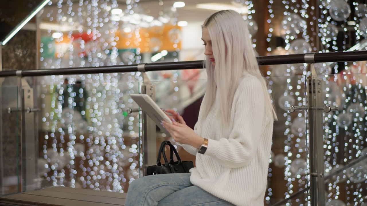 stylish shopper tapping tablet while seated on bench at decorated mall walkway, festive lights backdrop, casual fashion tech browsing moment in mall interior showcasing relaxed digital leisure style
