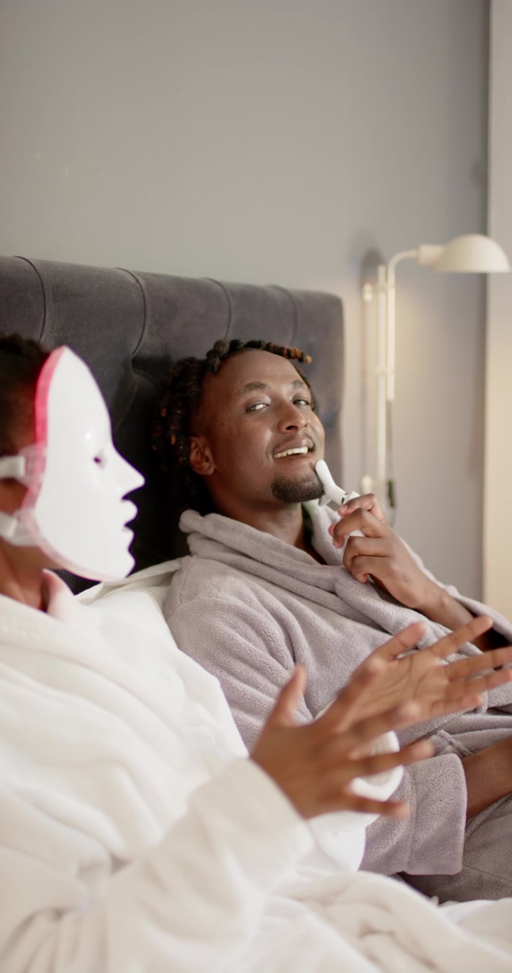 Vertical video: LED mask glowing is making couple use device, moving along jaw in bed for skincare