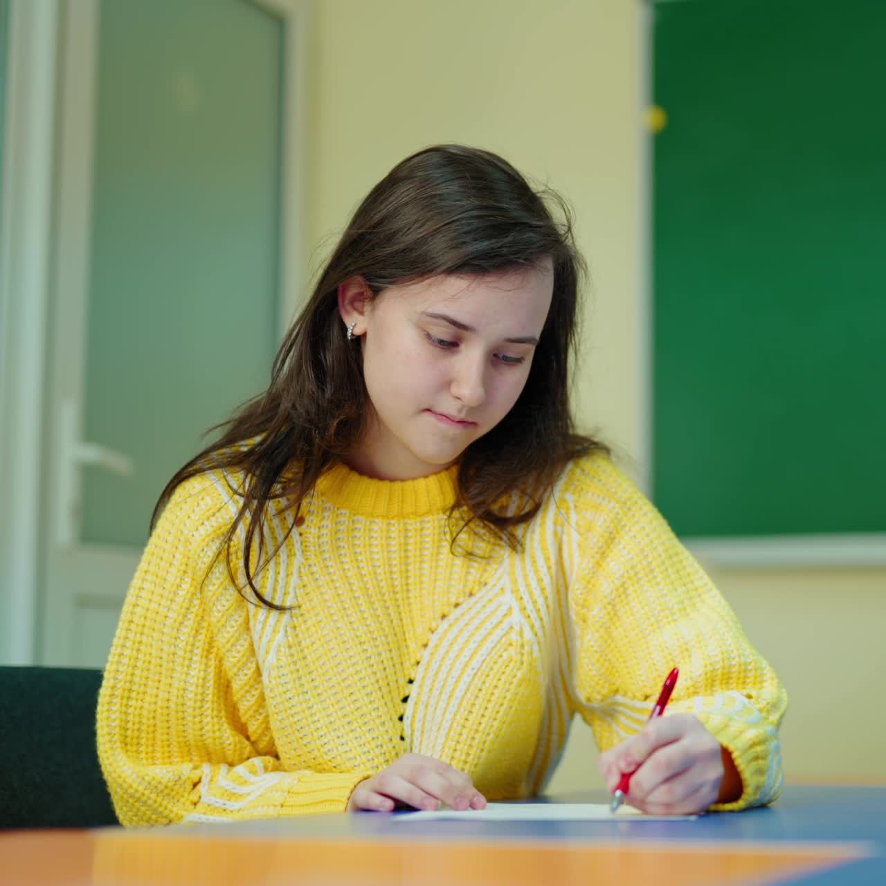 Thoughtful school girl writing at school. Portrait of a beautiful female student sitting at the desk and studying on the classroom background. Education at school.