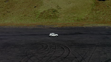 aerial: side follow of white car driving on black sand beach near green moss in iceland