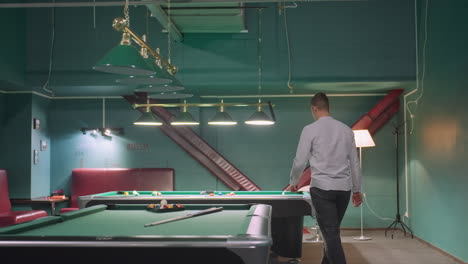 back shot of young man in white shirt and grey trousers walking into billiard room with modern interior, approaching pool table, and picking up cue stick. overhead lamps highlight gaming atmosphere