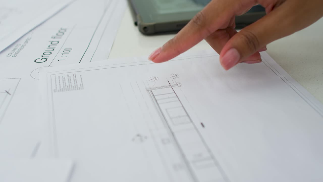 Placing finger on Ground Floor drawing, woman tracing, reviewing layout with phone near desk
