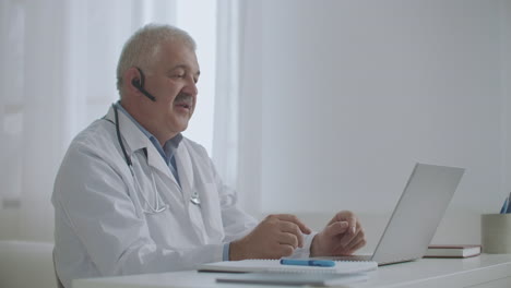 male therapist is sitting in his office of medical clinic and talking online by videochat with patients or colleagues