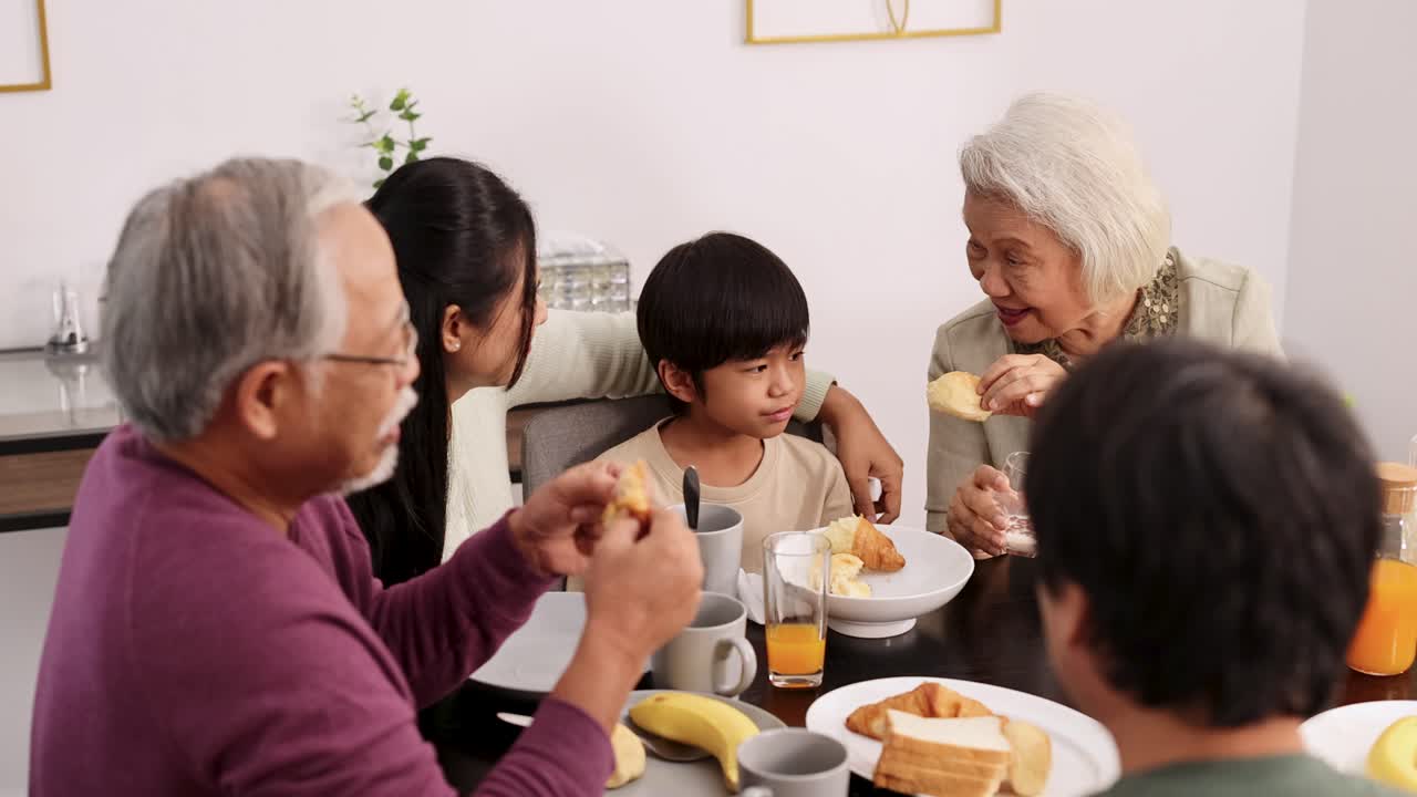 Three generations share a breakfast meal in a bright, modern dining room. Natural lighting, warm atmosphere, and candid interactions highlight family bonding