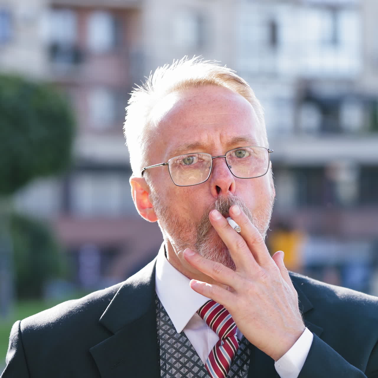Senior man in suit and tie with cigarette looks at camera outdoors. Portrait of a mature businessman with gray hair and eyeglasses smoking in the street.