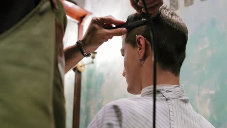 Young-bearded-barber-standing-and-making-stylish-haircut-of-attractive-man-with-clipper-in-barber-shot.-Bearded-man-is-sitting-on