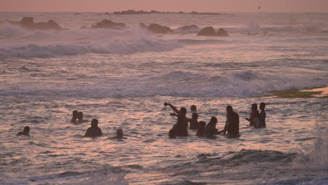 people splash in the ocean at sunset during the golden hour. the sky is painted pink. waves crash on the shore