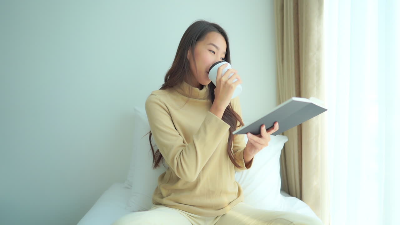 아늑한 거실 소파에서 재미있는 책을 읽고 흰색 컵에서 뜨거운 차나 커피를 마시는 젊은 아시아 매력적인 여성