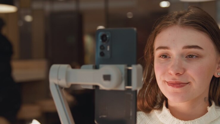 Giovane donna seduta dietro un gimbal portatile con uno smartphone montato in primo piano, sorride verso il dispositivo mentre lo stabilizzatore tiene fermo il telefono in una calda luce interna, suggerendo una configurazione da creator moderno, riprese.