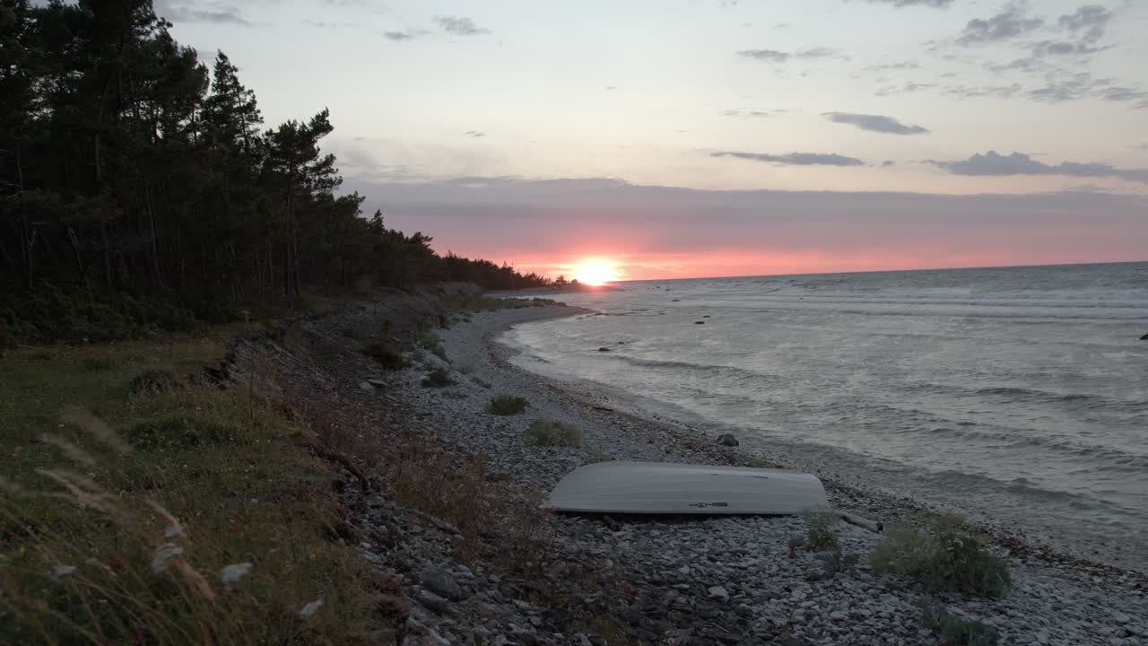 Baltic seashore during sunset, waves hitting the rocky beach with a small rowboat on the rocks, wind moving plants and trees